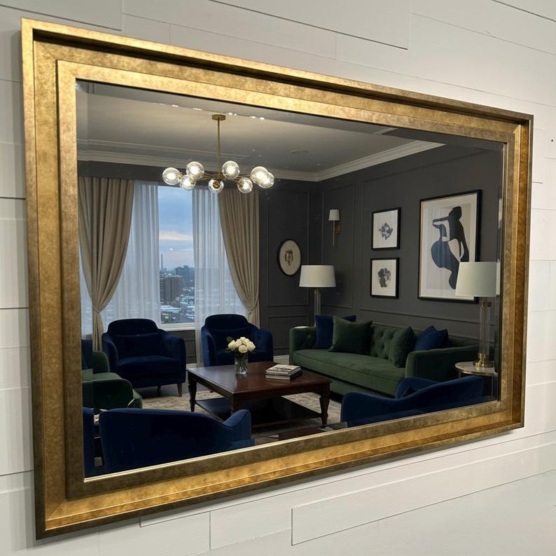 May include: A large rectangular wall mirror featuring a wide, textured gold frame with a distressed finish. The mirror reflects a living room interior containing a green sofa, dark blue armchairs, a wooden coffee table, and a chandelier.