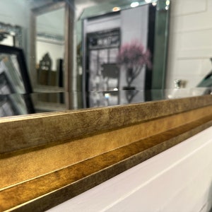 May include: A rectangular wall mirror featuring a wide, beveled frame with a distressed, antique gold finish. The frame has a multi-tiered, stepped profile that adds depth and texture to the edges, surrounding a clear, reflective glass surface.