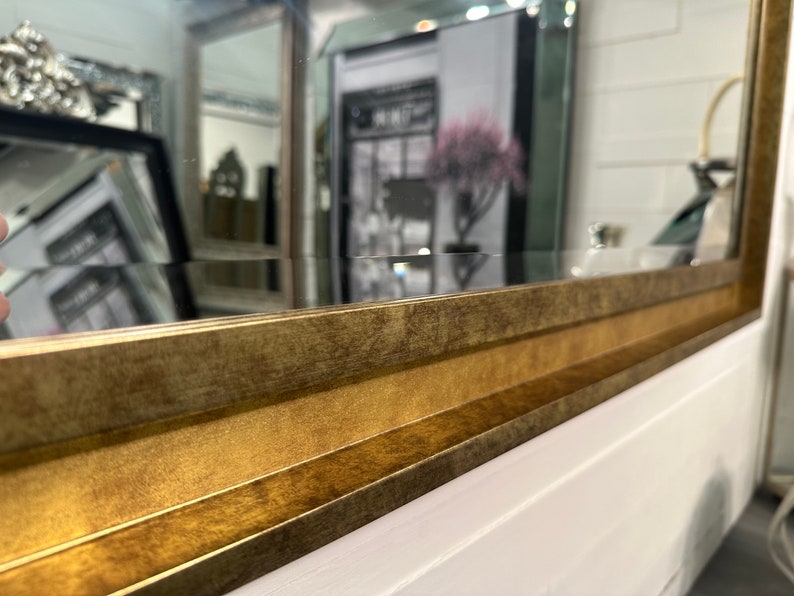 May include: A rectangular wall mirror featuring a wide, beveled frame with a distressed, antique gold finish. The frame has a multi-tiered, stepped profile that adds depth and texture to the edges, surrounding a clear, reflective glass surface.