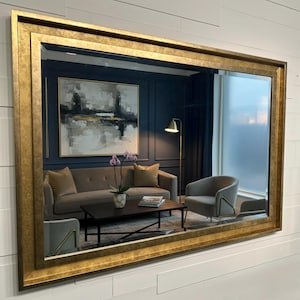 May include: A large rectangular wall mirror featuring a wide, textured gold frame with a distressed finish. The mirror reflects a living room interior containing a gray sofa, a dark coffee table, a floor lamp, and an abstract wall painting.