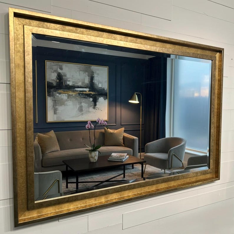 May include: A large rectangular wall mirror featuring a wide, textured gold frame with a distressed finish. The mirror reflects a living room interior containing a gray sofa, a dark coffee table, a floor lamp, and an abstract wall painting.