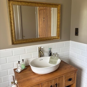 May include: A bathroom vanity featuring a wooden cabinet with black hardware, a white oval vessel sink, and a brass faucet. Above the sink is a rectangular mirror with a gold frame, mounted on a wall with white subway tiles and tan paint.