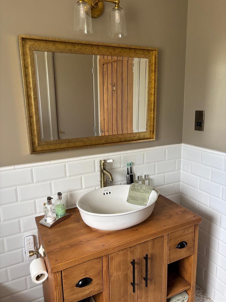 May include: A bathroom vanity featuring a wooden cabinet with black hardware, a white oval vessel sink, and a brass faucet. Above the sink is a rectangular mirror with a gold frame, mounted on a wall with white subway tiles and tan paint.