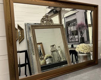Op de afbeelding: Een grote rechthoekige spiegel met een bronskleurige lijst. De spiegel reflecteert andere spiegels en decoratieve items, waaronder een zilveren decoratief object, vazen en een zwart-wit afbeelding van een winkel. De lijst heeft een dubbellaags ontwerp.