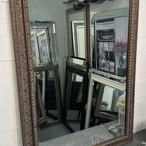 May include: A large, ornate, gold-toned mirror with a decorative frame. The frame has a raised, textured design with a rope-like border. The mirror is reflecting other mirrors and a sign that says "Dior".