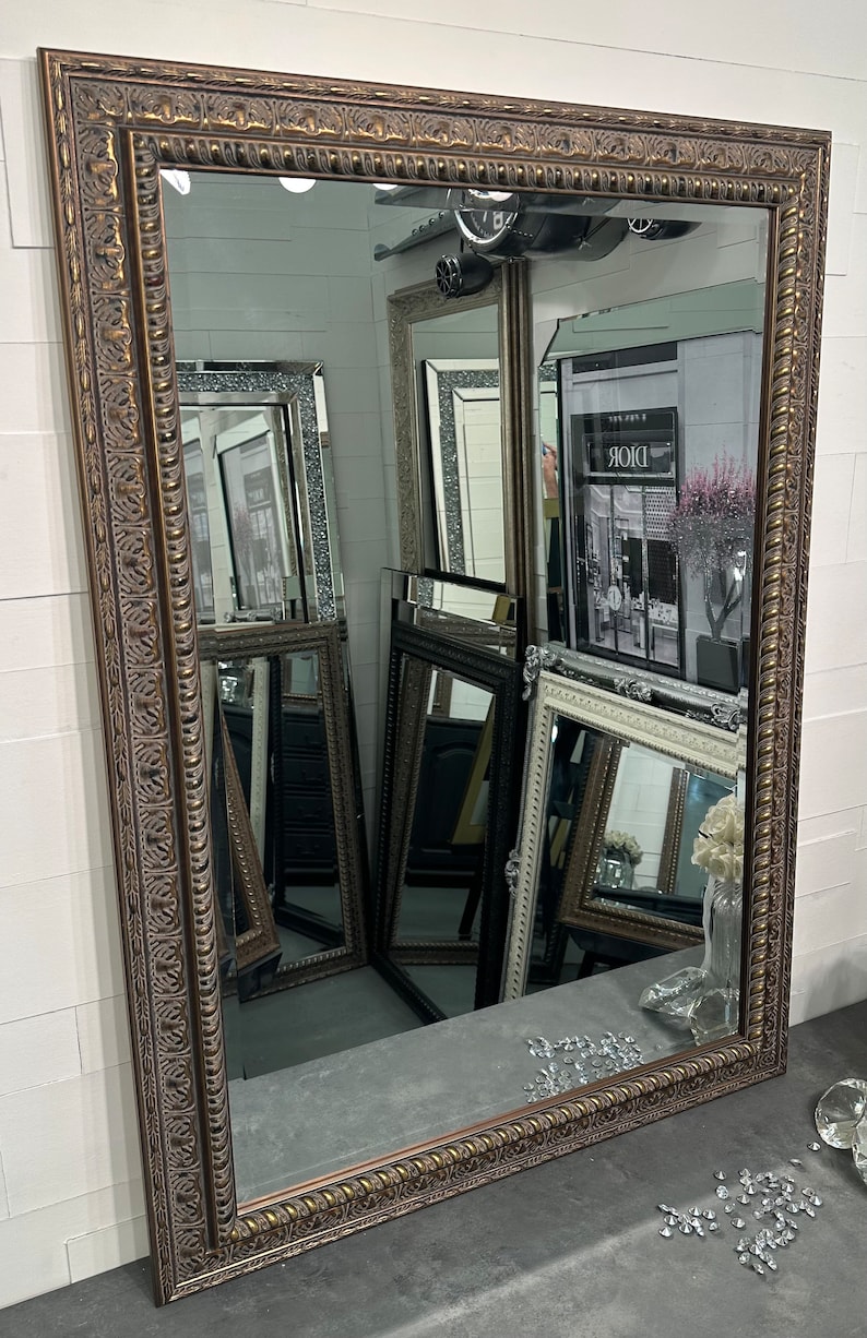 May include: A large, ornate, gold-toned mirror with a decorative frame. The frame has a raised, textured design with a rope-like border. The mirror is reflecting other mirrors and a sign that says "Dior".
