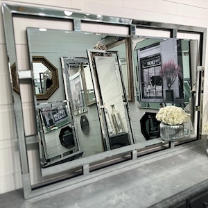 May include: A large rectangular mirror with a silver frame and beveled edges. The mirror reflects several other mirrors of various shapes and sizes, along with a shop front and a vase of white flowers. The overall aesthetic is elegant and decorative.