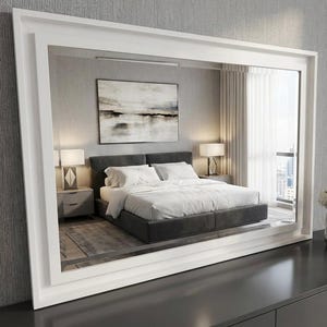 May include: A large, rectangular white framed mirror reflecting a modern bedroom. The bedroom features a grey bed with white bedding, two bedside tables with lamps, and a painting on the wall. The mirror is on a dark grey surface.