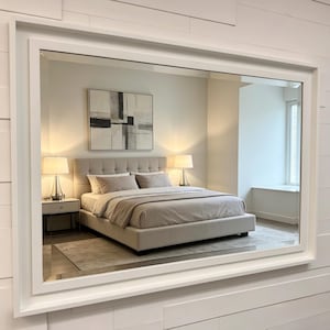 May include: A large rectangular wall mirror with a thick white frame. The mirror reflects a bedroom scene featuring a tufted beige bed, two bedside tables with lamps, and an abstract black, white, and grey painting hanging on the wall.