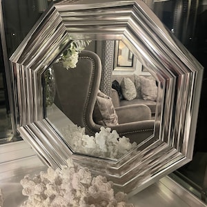 May include: Octagonal silver-toned mirror with a bevelled edge and a decorative design. The mirror is reflecting a room with a chair and a sofa.