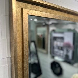 May include: A rectangular wall mirror featuring a thick, textured gold frame with a stepped, multi-layered profile. The frame has a matte, metallic finish with subtle variations in tone, creating a deep, dimensional appearance around the glass.