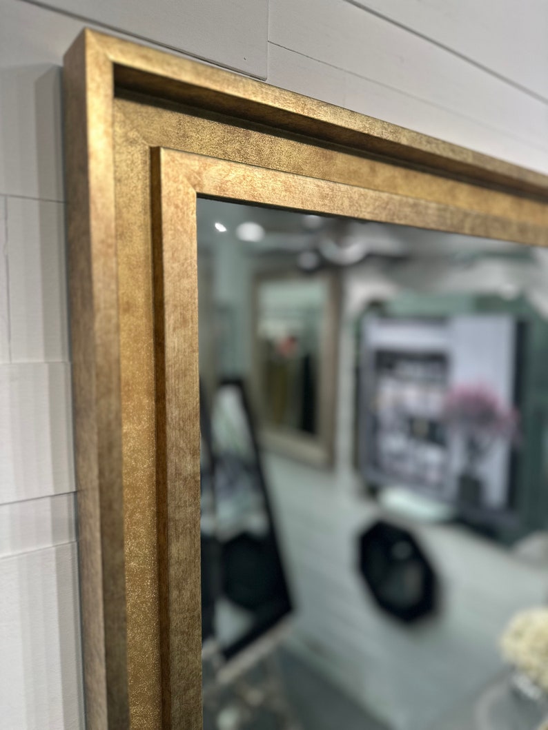 May include: A rectangular wall mirror featuring a thick, textured gold frame with a stepped, multi-layered profile. The frame has a matte, metallic finish with subtle variations in tone, creating a deep, dimensional appearance around the glass.