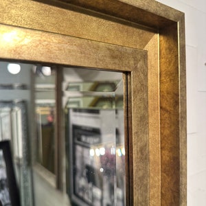 May include: A rectangular wall mirror featuring a wide, textured gold-toned frame with a beveled edge. The frame has a matte, metallic finish with subtle variations in color, and the glass surface shows a clear, angled bevel along the inner border.