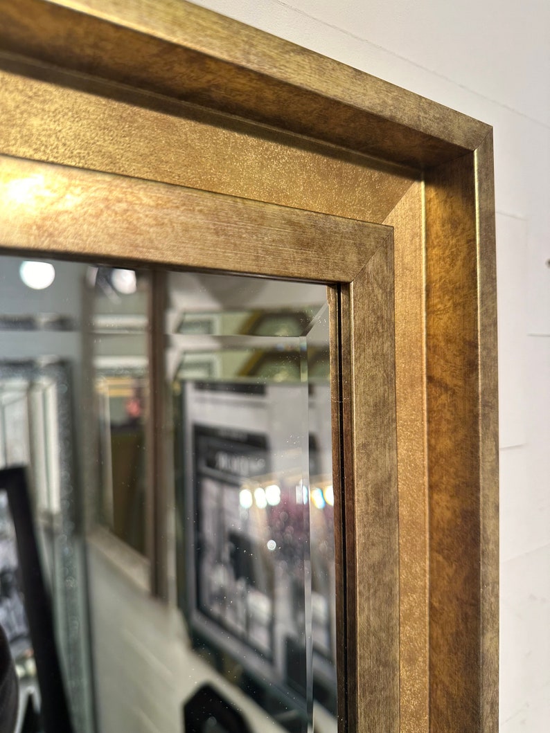 May include: A rectangular wall mirror featuring a wide, textured gold-toned frame with a beveled edge. The frame has a matte, metallic finish with subtle variations in color, and the glass surface shows a clear, angled bevel along the inner border.