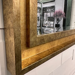 May include: A rectangular wall mirror featuring a wide, textured frame with a metallic gold finish. The frame has a multi-layered, stepped profile with a distressed appearance. The reflective glass surface shows a partial reflection of a storefront.