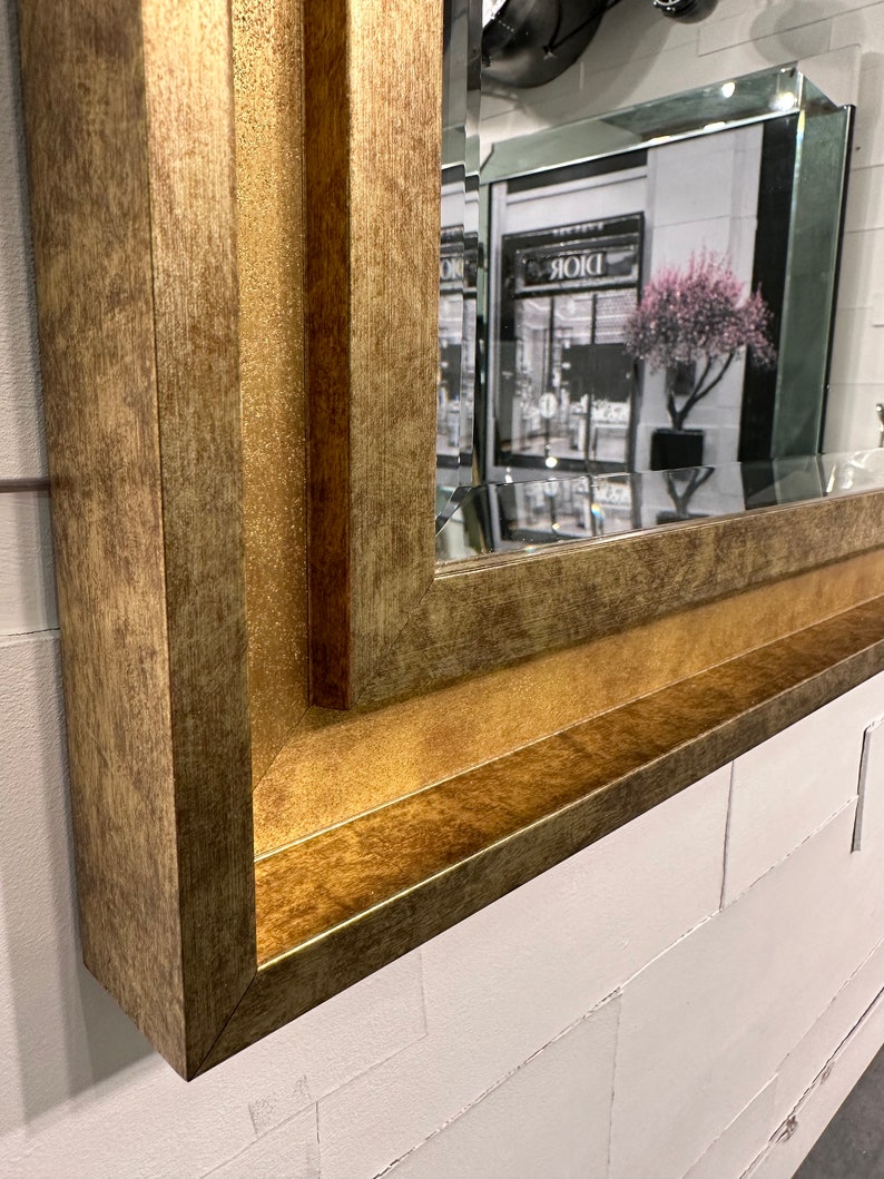 May include: A rectangular wall mirror featuring a wide, textured frame with a metallic gold finish. The frame has a multi-layered, stepped profile with a distressed appearance. The reflective glass surface shows a partial reflection of a storefront.
