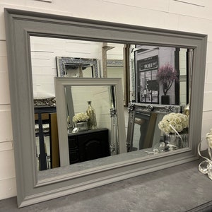 May include: A large grey framed mirror with a bevelled edge. The mirror reflects other mirrors and a shop front with the word "Dior" visible.