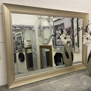 May include: A silver framed mirror with a bevelled edge and a decorative design. The mirror is reflecting other mirrors and a vase with white flowers.