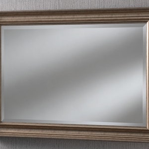 May include: A rectangular wall mirror featuring a wide, metallic bronze-toned frame with a stepped, beveled profile. The reflective glass surface is clear and smooth, set within the ornate, multi-layered border that adds depth to the design.