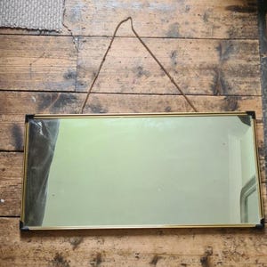 Puede incluir: Un espejo de pared rectangular con un marco de metal fino en tono dorado y detalles negros en las esquinas. El espejo está suspendido por una cadena de metal fina de color dorado sujeta al marco superior, permitiendo colgarlo vertical u horizontalmente.