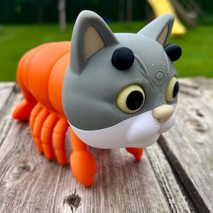 Könnte beinhalten: Eine skurrile 3D-gedruckte Skulptur eines Katze-Hummer-Hybriden. Der Katzenkopf ist grau mit schwarzen Akzenten und großen gelben Augen. Der Körper ist orange, wie ein Hummer. Auf einer verwitterten Holzoberfläche präsentiert.