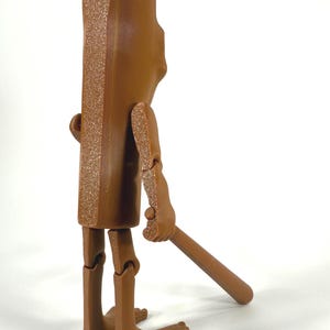 May include: A brown articulated figure with a rectangular body and a textured edge. The figure has jointed arms and legs, and is holding a brown stick. The figure is standing on a white surface.