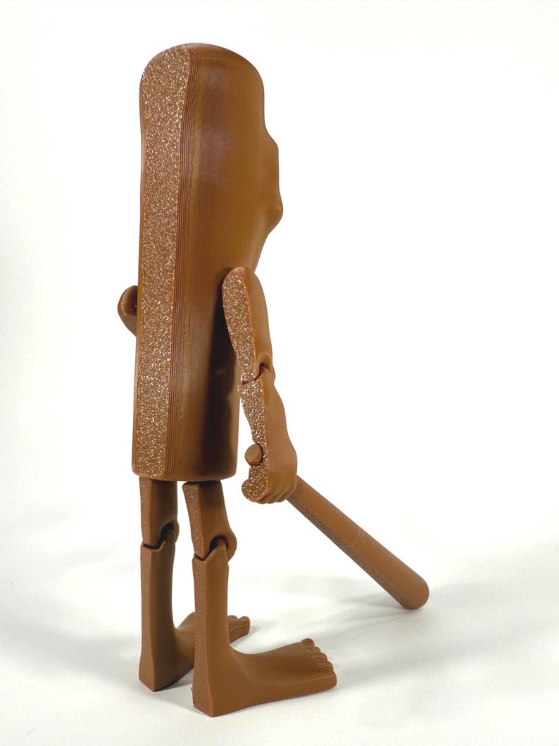 May include: A brown articulated figure with a rectangular body and a textured edge. The figure has jointed arms and legs, and is holding a brown stick. The figure is standing on a white surface.