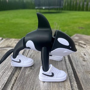 Puede incluir: Una figura de orca en blanco y negro con zapatillas blancas con swooshes negros. La orca tiene una aleta dorsal y manchas blancas. La figura está sobre una superficie de madera con un fondo verde.