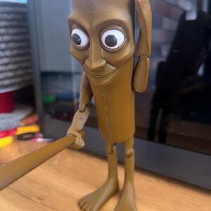 May include: A brown, articulated figurine with large white-eyed features, holding a brown bat. The figure has a rectangular head, a slender body, and bare feet. The figure is posed with one hand on its head.