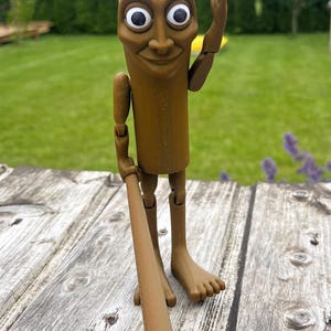 May include: A brown, articulated figurine with a rectangular body, large white-eyed face, and a smiling expression. It holds a brown baseball bat and is posed with one arm raised. The figurine stands on a weathered wooden surface.