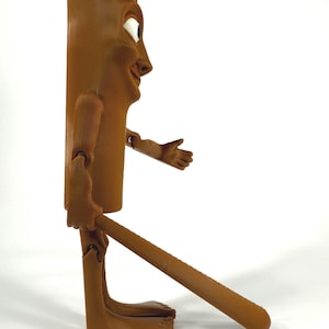 May include: A brown, articulated wooden figure with a smiling face, outstretched arm, and a long, cylindrical body. The figure has a whimsical design, with a long leg extending from the body. The figure is standing on a white surface.