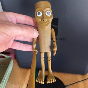 May include: A tan-coloured, articulated figurine with a cylindrical body, large eyes, and a smiling expression. The figurine is holding a small baseball bat. The figure has jointed arms and legs, and is standing on a flat surface.
