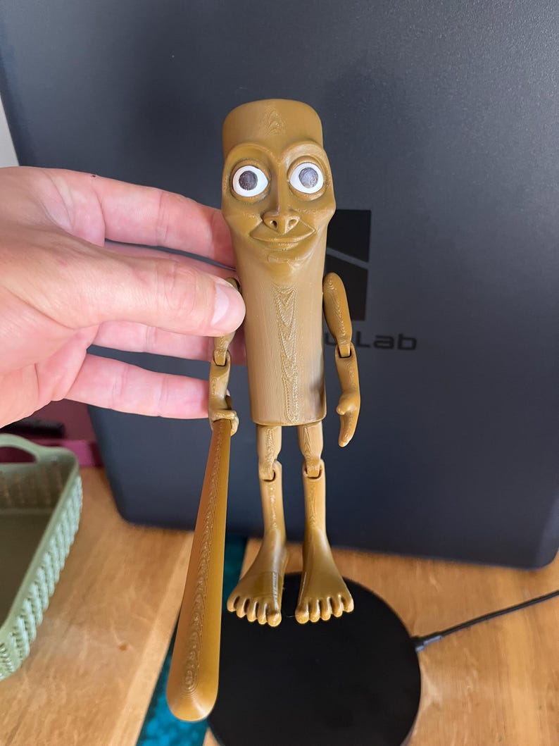 May include: A tan-coloured, articulated figurine with a cylindrical body, large eyes, and a smiling expression. The figurine is holding a small baseball bat. The figure has jointed arms and legs, and is standing on a flat surface.