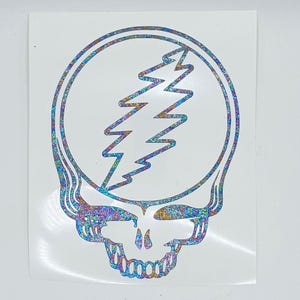 May include: A holographic sticker featuring a stylized skull design with a lightning bolt centered within a circular outline. The graphic has a reflective, multi-colored glitter finish that shifts in appearance against a plain white background.