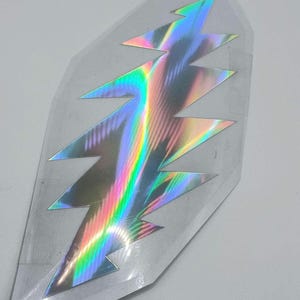 May include: A holographic sticker featuring a lightning bolt design in a silver, pink, and blue color scheme.