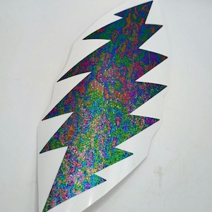 May include: A white, die-cut sticker in the shape of a lightning bolt. The interior of the bolt has a colorful, iridescent pattern with hues of green, blue, purple, and gold. The sticker is on a white background.