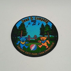 May include: Circular sticker featuring the text "Dead & Company 2025" with a scene of dancing bears, the Golden Gate Bridge, and musical notes. The bears are blue and orange, set against a green and blue background. The text "Golden Gate Park San Francisco" is also included.