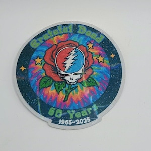 May include: A round sticker featuring the Grateful Dead logo: a red rose with a blue and red lightning bolt skull. The sticker has a tie-dye background and the text "Grateful Dead" and "60 Years 1965-2025" in a circular pattern.