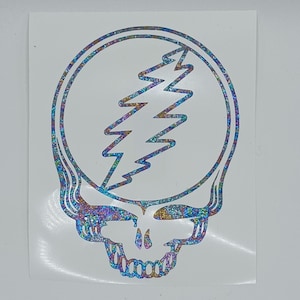 May include: A holographic sticker featuring a skull with a lightning bolt. The skull is outlined in a shimmering, iridescent material, with the lightning bolt in the center. The design is on a white background.