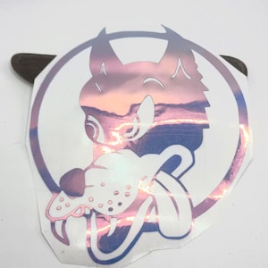 May include: A circular holographic sticker featuring a stylized wolf head graphic with a prominent tongue and pointed ears. The surface displays a shifting metallic gradient of pink, purple, and blue tones, creating a reflective effect within the design.