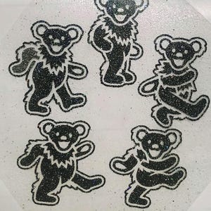 May include: A set of five black glittery bear-shaped decals arranged in a circular pattern. Each bear is depicted in a dancing pose with a white outline, a smiling face, and a ruff of fur around the neck, all set against a clear, shimmering background.