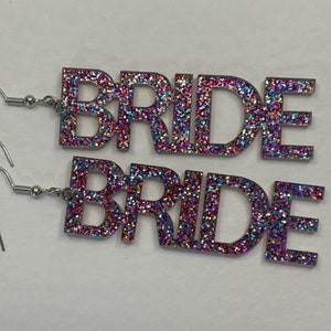 Op de afbeelding: Een paar zilveren oorbellen met het woord "BRIDE" gespeld in glinsterende roze, blauwe en paarse letters.
