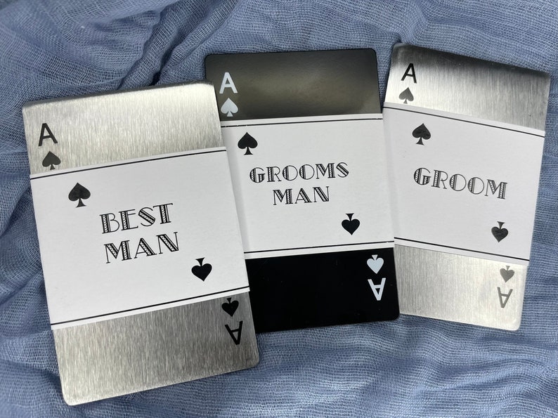 Może przedstawiać: Trzy srebrne metalowe karty z czarnym tekstem i czarnym wzorem pik. Karty brzmią "Best Man", "Grooms Man" i "Groom".