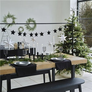 May include: A black and white star garland hangs above a table set for a festive dinner. The table is decorated with black and white plates, black napkins, and candles. A Christmas tree with white lights is in the background.