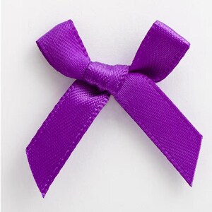 Peut inclure: Un petit nœud en satin violet vif. Le nœud est noué au centre avec deux boucles et deux rubans. Le tissu a un léger éclat et le nœud est posé sur un fond blanc.