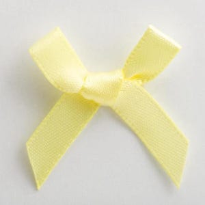 Pode incluir: Uma pequena fita de cetim amarelo pálido. A fita é amarrada no centro com duas voltas e duas fitas. A fita é feita de um tecido liso e brilhante. A fita está em um fundo branco.