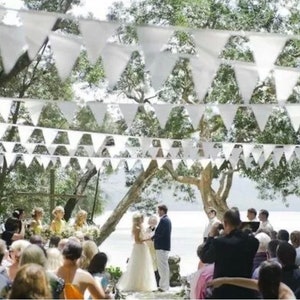 May include: Outdoor wedding ceremony with white triangular bunting strung overhead. A couple stands at the altar, surrounded by guests. The scene is set under a canopy of trees, creating a natural and festive atmosphere.