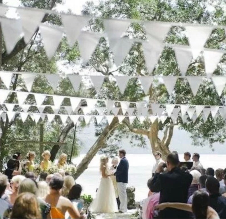 May include: Outdoor wedding ceremony with white triangular bunting strung overhead. A couple stands at the altar, surrounded by guests. The scene is set under a canopy of trees, creating a natural and festive atmosphere.
