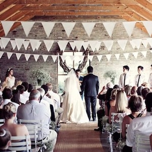 May include: A wedding ceremony is taking place in a rustic venue. White triangular pennant banners hang from the wooden ceiling. The bride and groom stand at the altar, facing the officiant, with guests seated on either side.