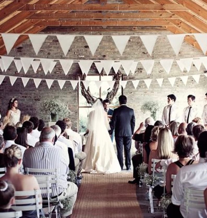 May include: A wedding ceremony is taking place in a rustic venue. White triangular pennant banners hang from the wooden ceiling. The bride and groom stand at the altar, facing the officiant, with guests seated on either side.